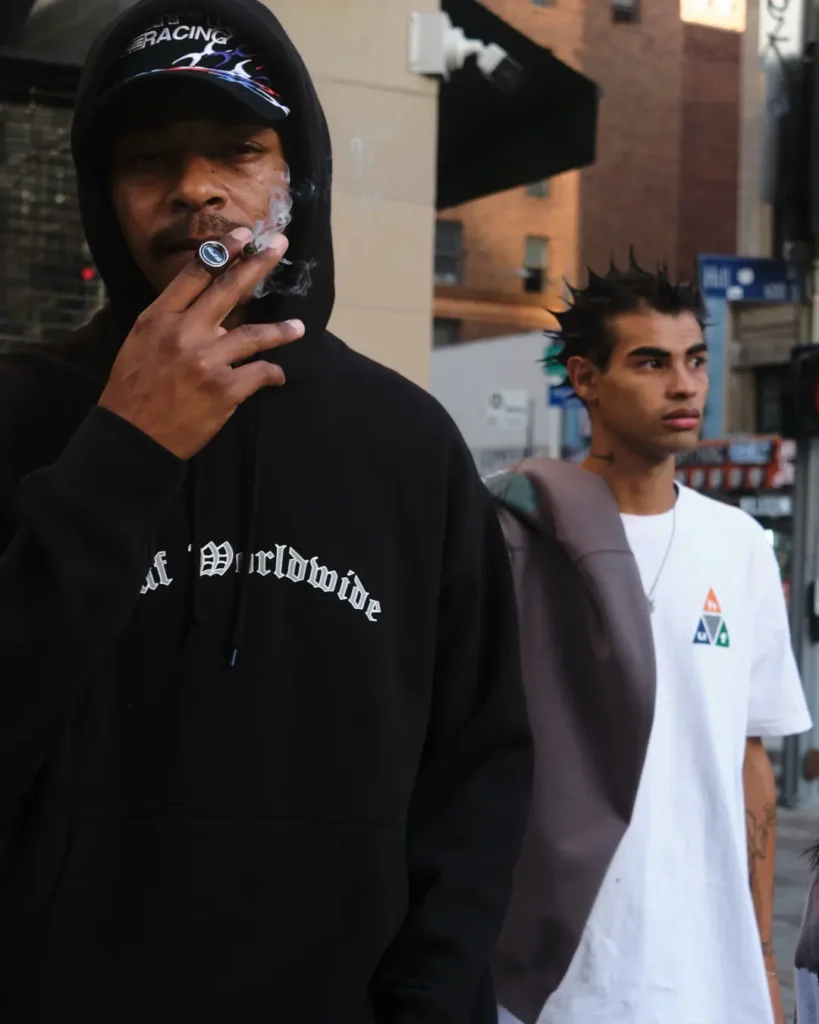 Two modelon a city street, one wear a black HUF hoodie and cap and another wear an oversize white HUF graphic Tee