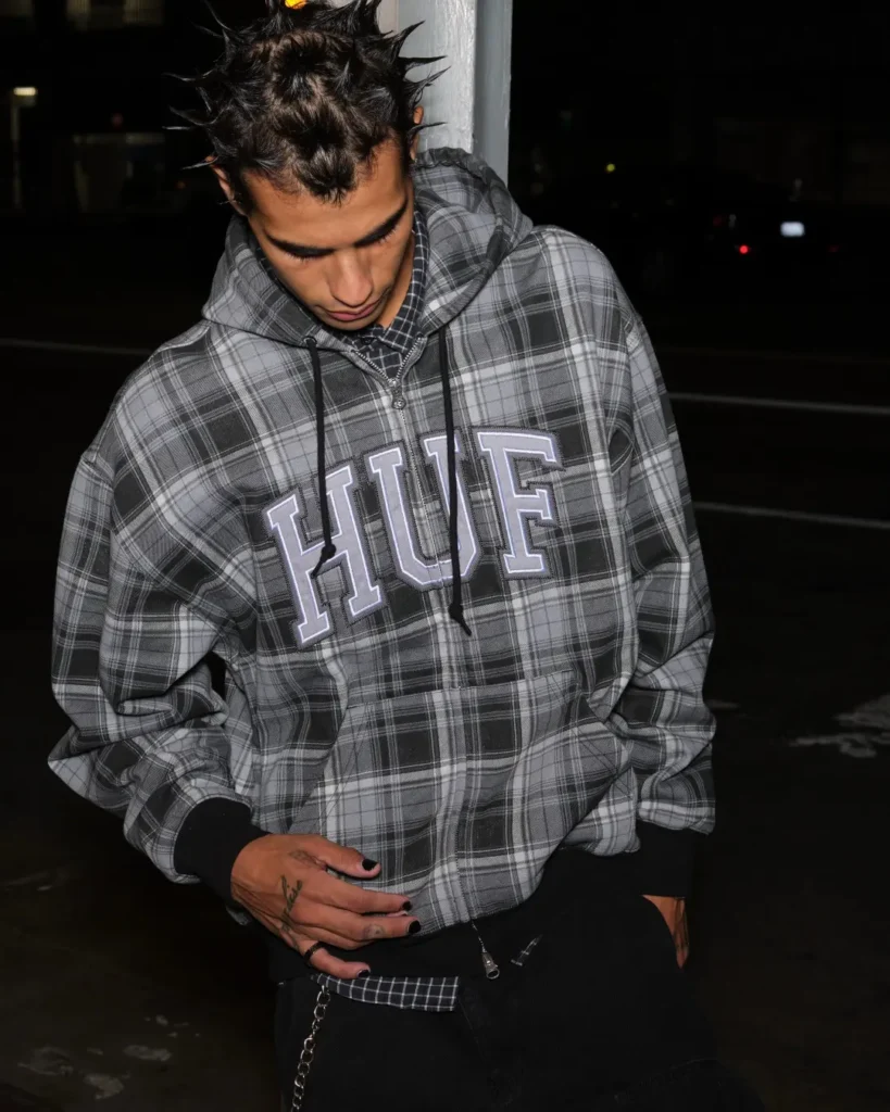 Model with spike hair wear an oversize gray plaid HUF zip-up hoodie with large collegiate-style letter