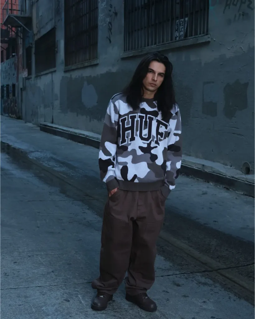 Long-hair model wearing a gray camouflage HUF sweater and loose brown trousers in a dim urban alley