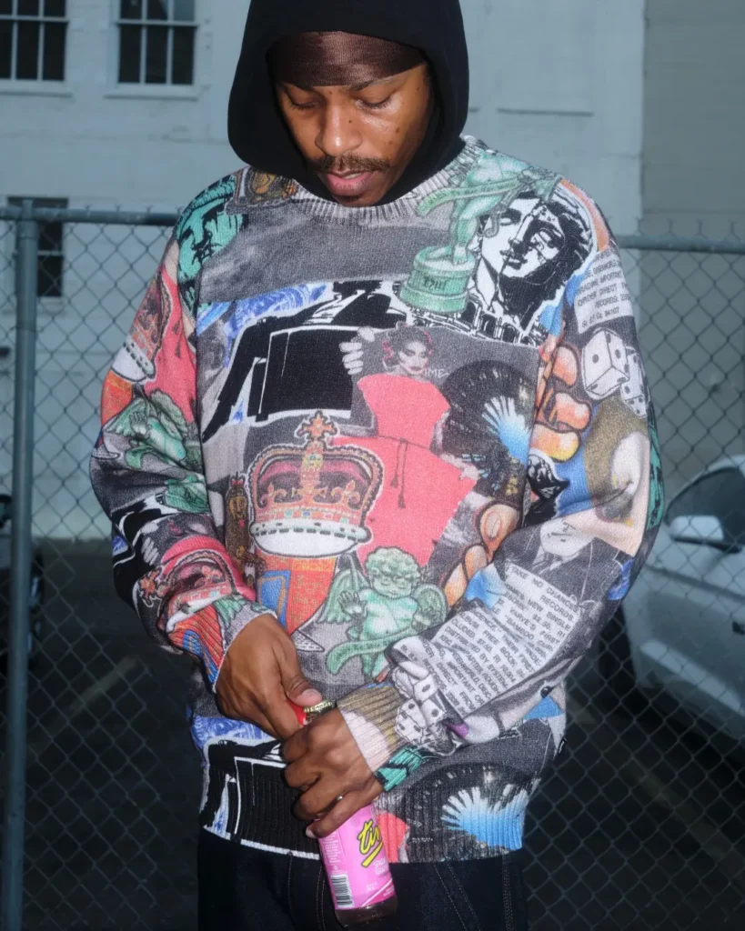 Model wear a colorful collage-print HUF sweater layered over a black hooded top beside a chain-link fence