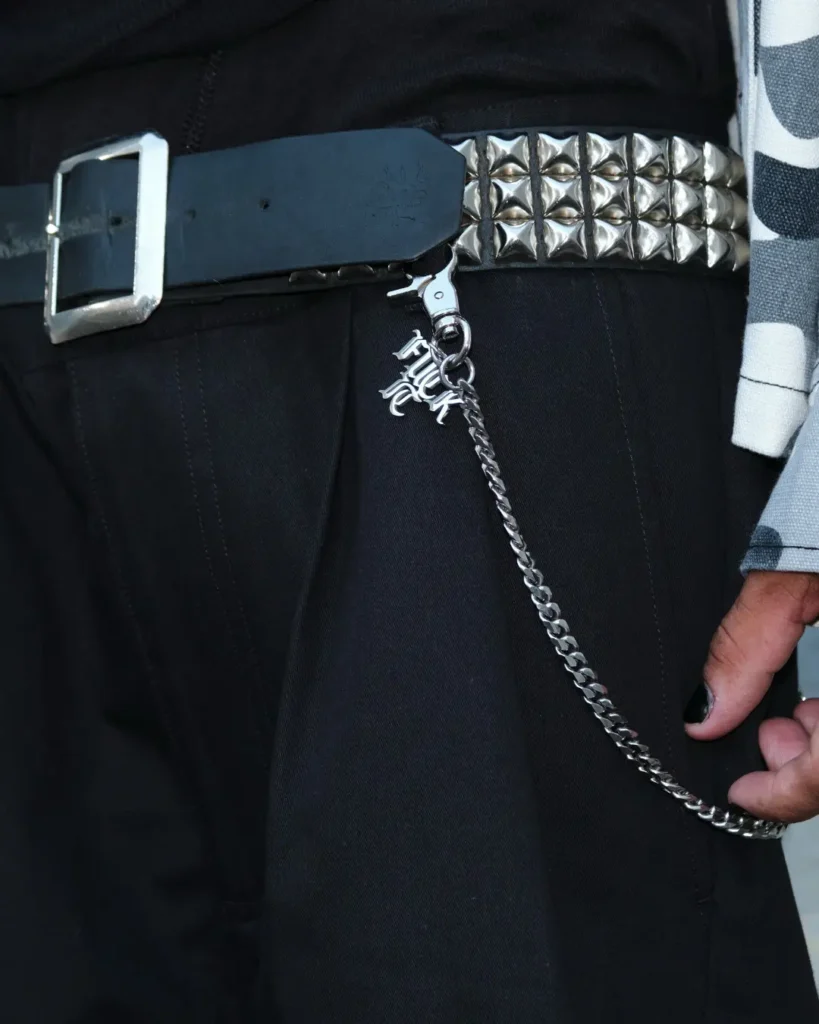 Close-up of black trousers styled with a silver pyramid-studded belt, metal wallet chain and HUF letter charm