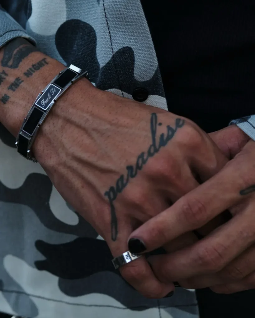 Close-up of tattoo hands with black-painte nails, a silver ring and black-and-silver bracelet against a gray camouflage HUF garment