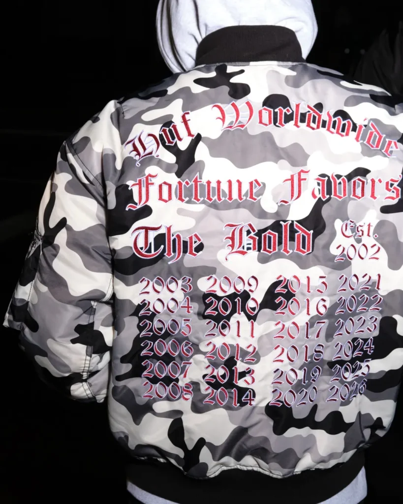 Close rear view of a gray camouflage HUF bomber jacket embroidered with “Huf Worldwide Fortune Favors The Bold” and years from 2003 through 2026