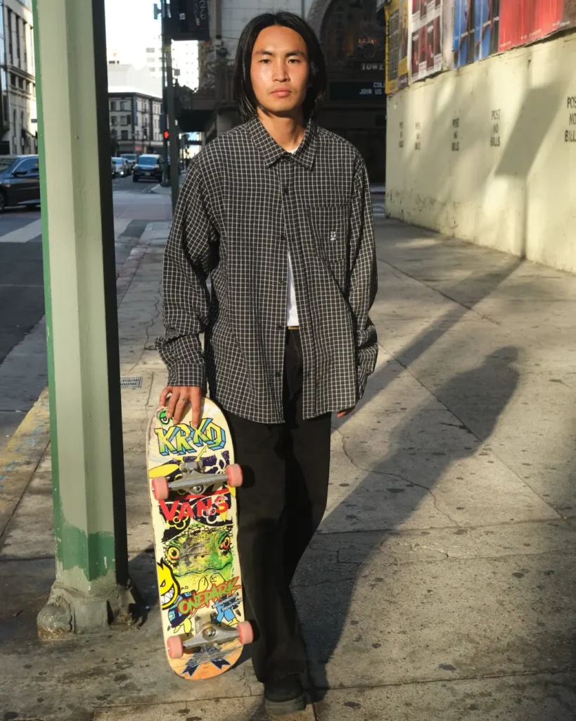 Model in an oversized black grid-pattern shirt and wide black trousers holding a graphic skateboard on a sunlit city sidewalk