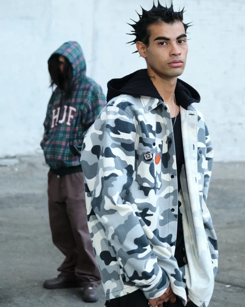 Model with spiked black hair wearing a gray camouflage HUF overshirt layered over a black hoodie, with a plaid HUF jacket view in the background