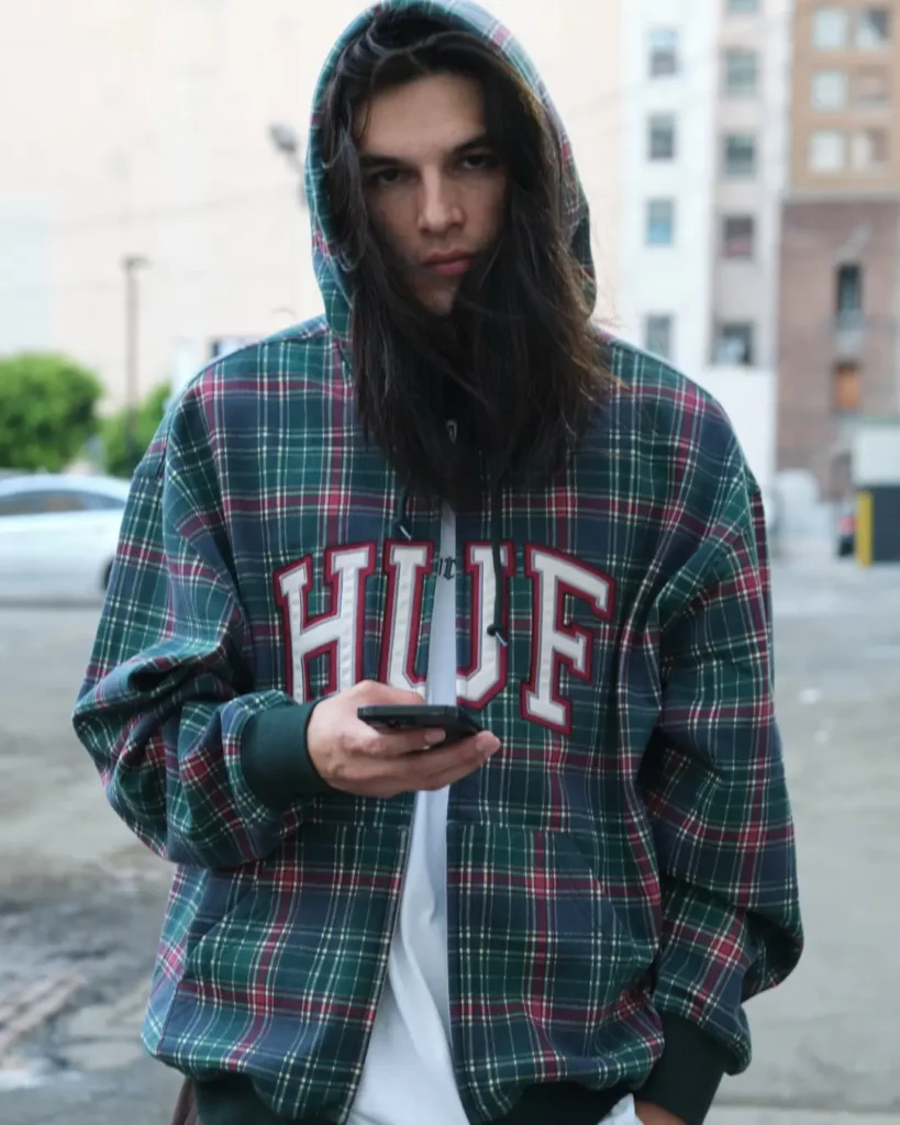 Long-haired model wear an oversize green, red and navy plaid HUF hooded jacket while hold a phone outdoor