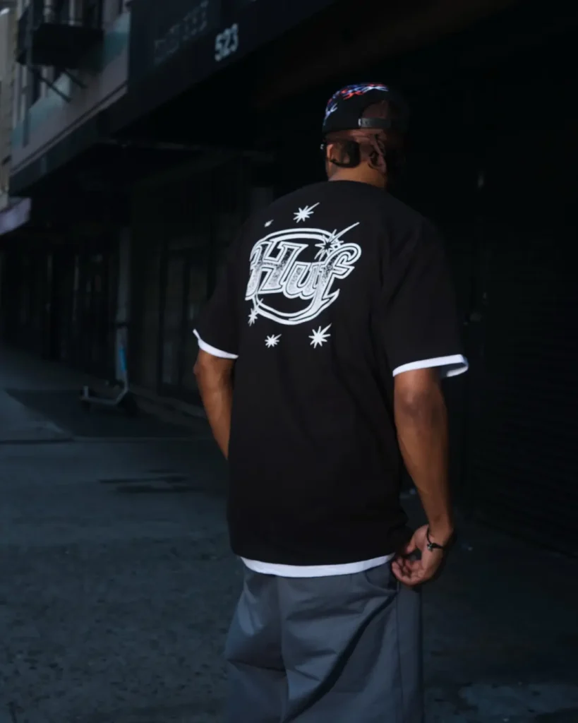 Rear view of a model wear a black HUF graphic Tee with a large white script logo and star illuminated