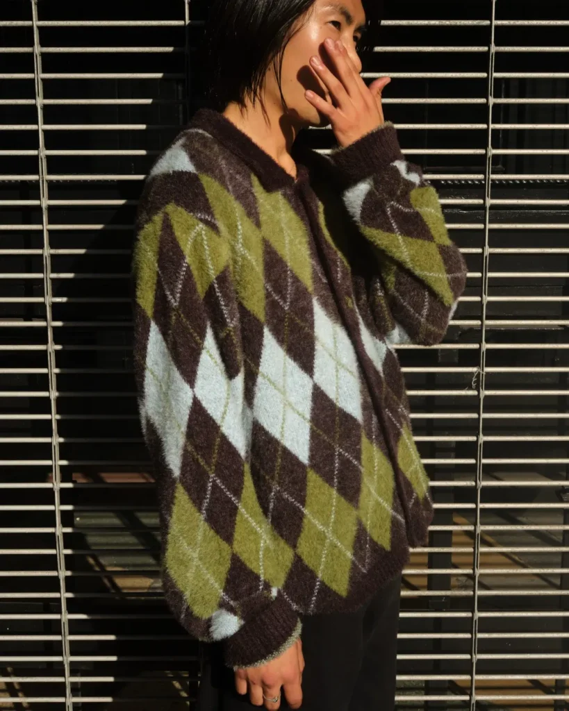 Model wear an oversize fuzzy argyle cardigan in brown, olive green and pale blue against a metal storefront gate