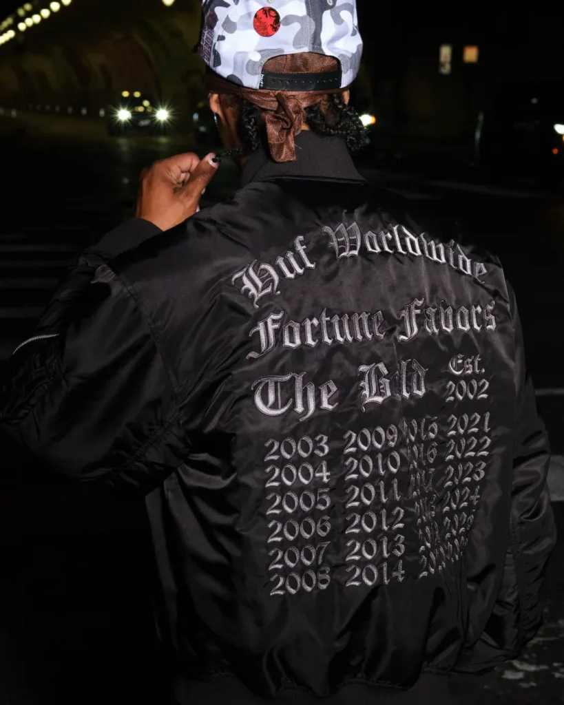 Rear view of a black HUF bomber jacket embroidered with “Huf Worldwide Fortune Favors The Bold” and a timeline of year