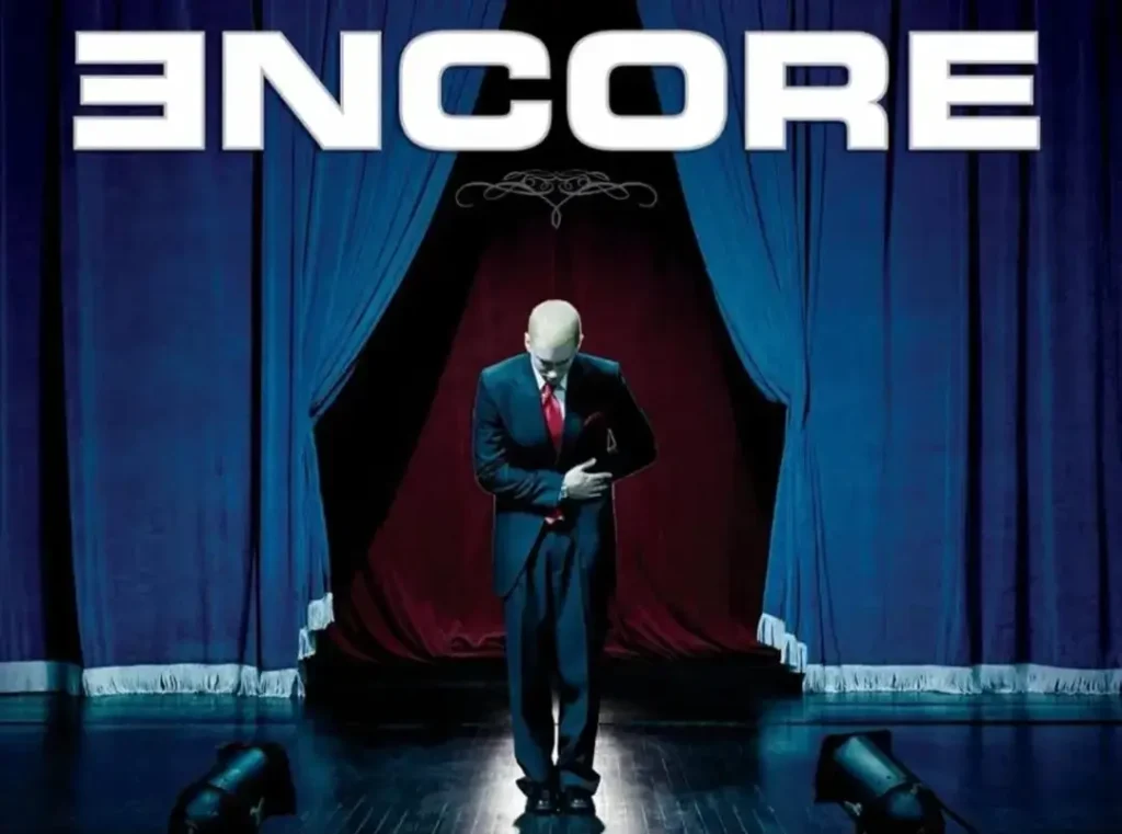 Eminem bows on a theatrical stage between blue curtains beneath large white “ENCORE” lettering, wearing a dark suit and red tie