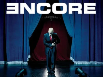 Eminem bows on a theatrical stage between blue curtains beneath large white “ENCORE” lettering, wearing a dark suit and red tie