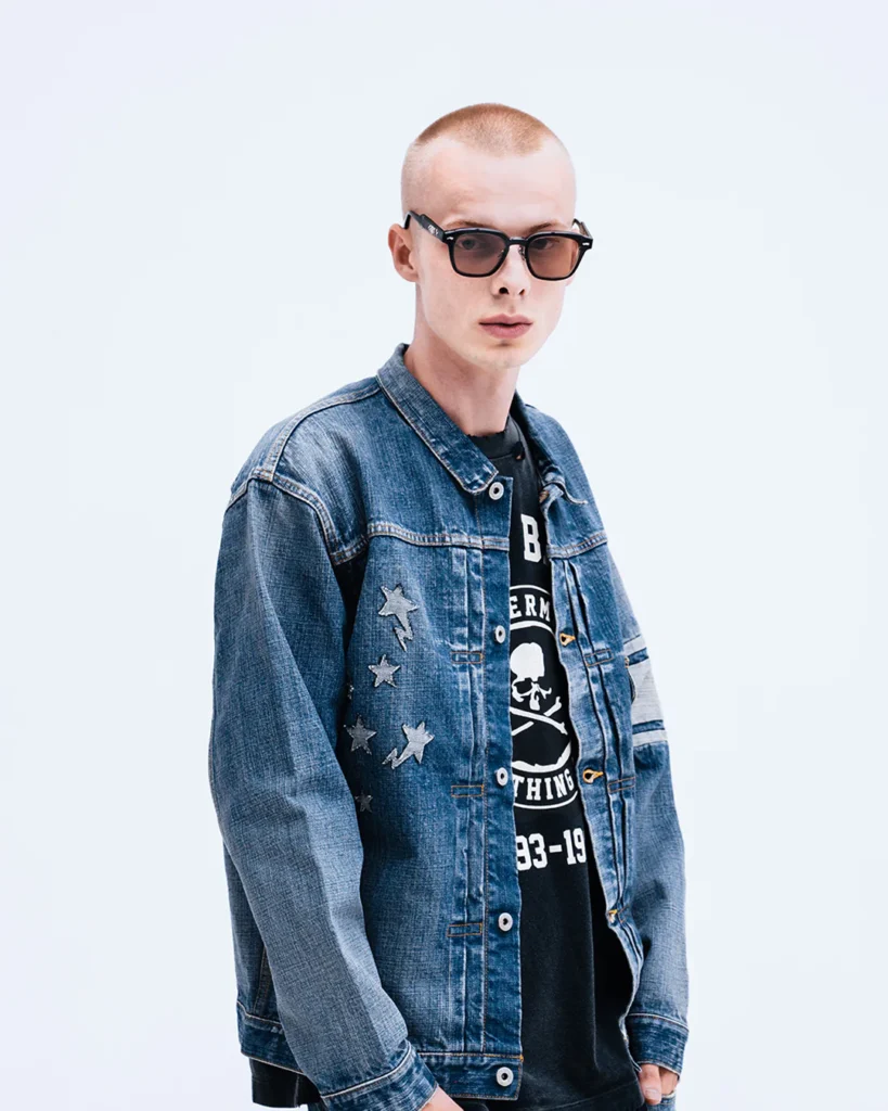 Model in sunglasses wearing a faded blue denim jacket with star appliqués over a black Mastermind x BAPE graphic shirt