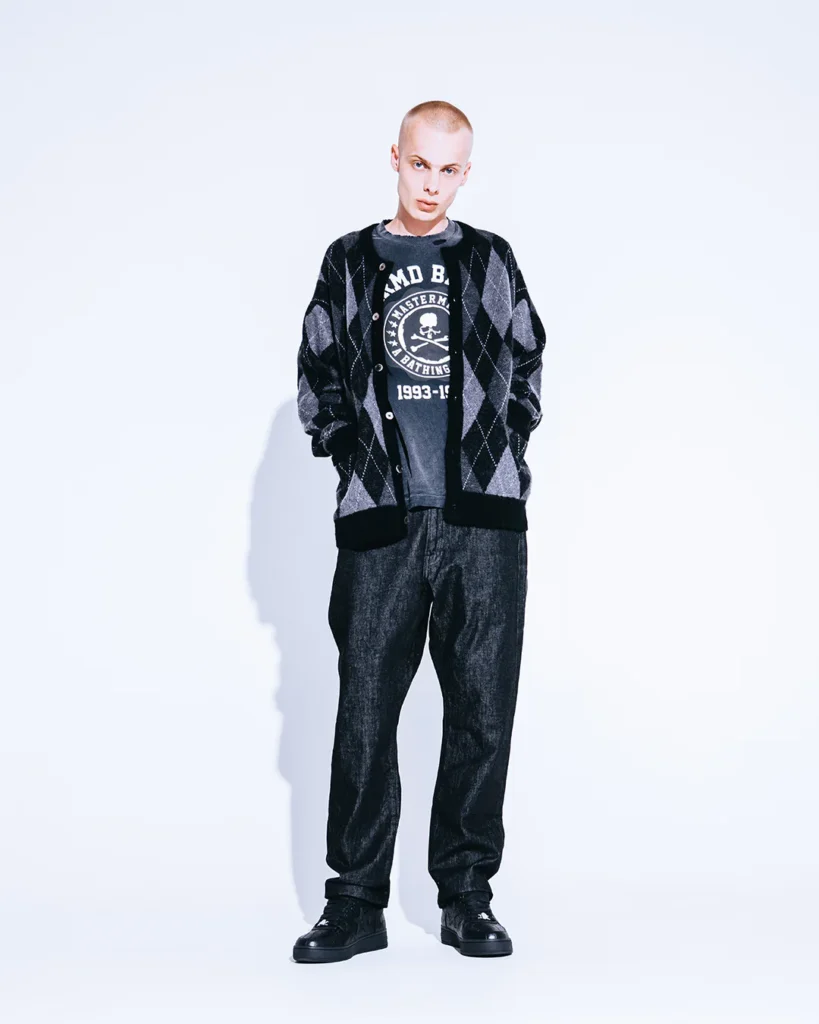 Full-length studio look with a black-and-gray argyle cardigan, Mastermind x BAPE graphic top, dark loose-fit jeans and black shoe