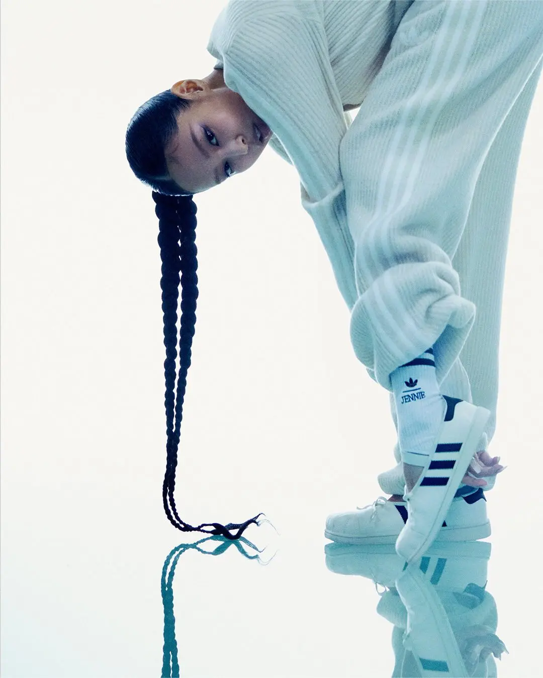 Jennie Bends Forward In A White Outfit While Holding A White Adidas Ballet Shoe Near A Reflective Floor.