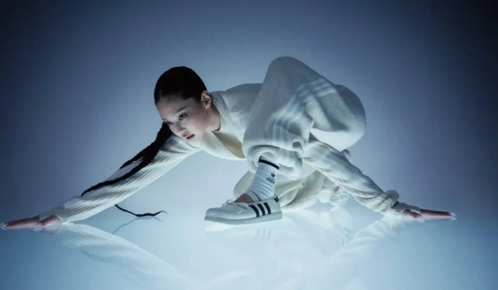 Model Poses Low To The Floor In A White Outfit While Wearing White Adidas Jennie Ballet Shoe