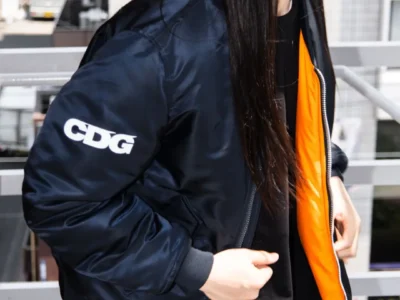 Black CDG MA-1 bomber jacket with a bright orange lining, worn over a black outfit, featuring a bold white CDG logo printed on the sleeve