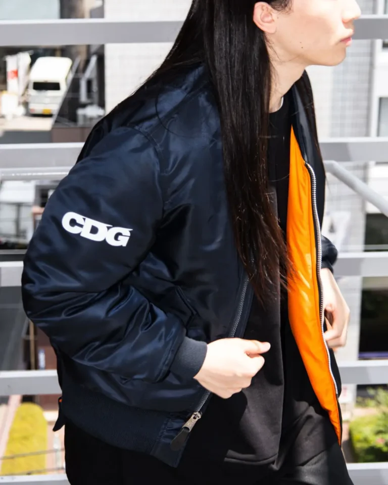 Black CDG MA-1 bomber jacket with a bright orange lining, worn over a black outfit, featuring a bold white CDG logo printed on the sleeve