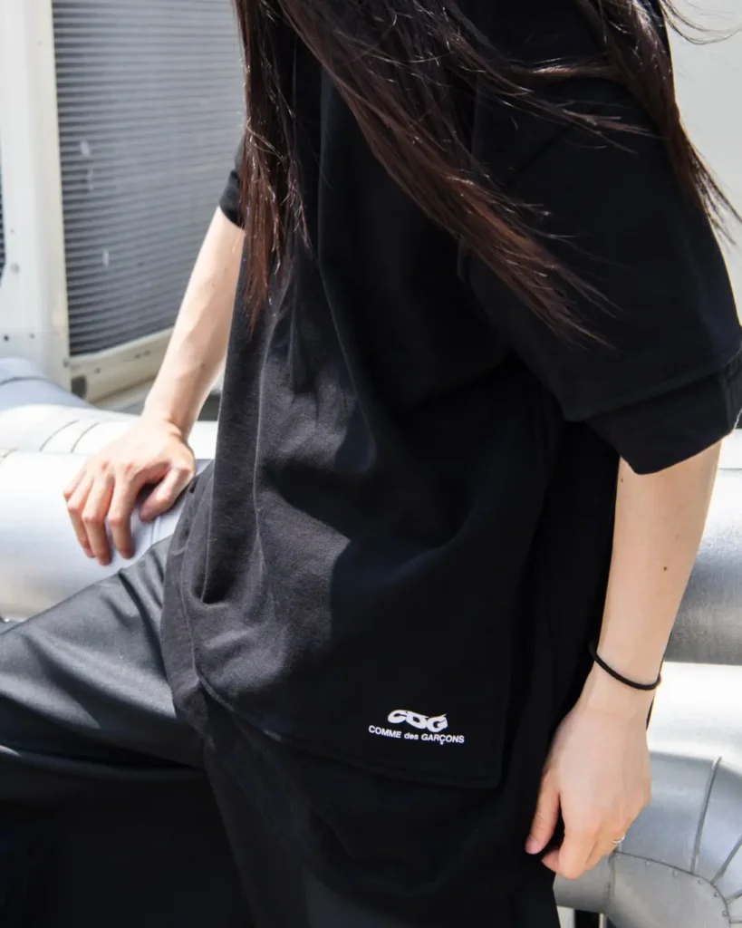 Oversized black CDG Tee with a small white COMME des GARÇONS logo near the hem, styled with relaxed black pants in an industrial outdoor set