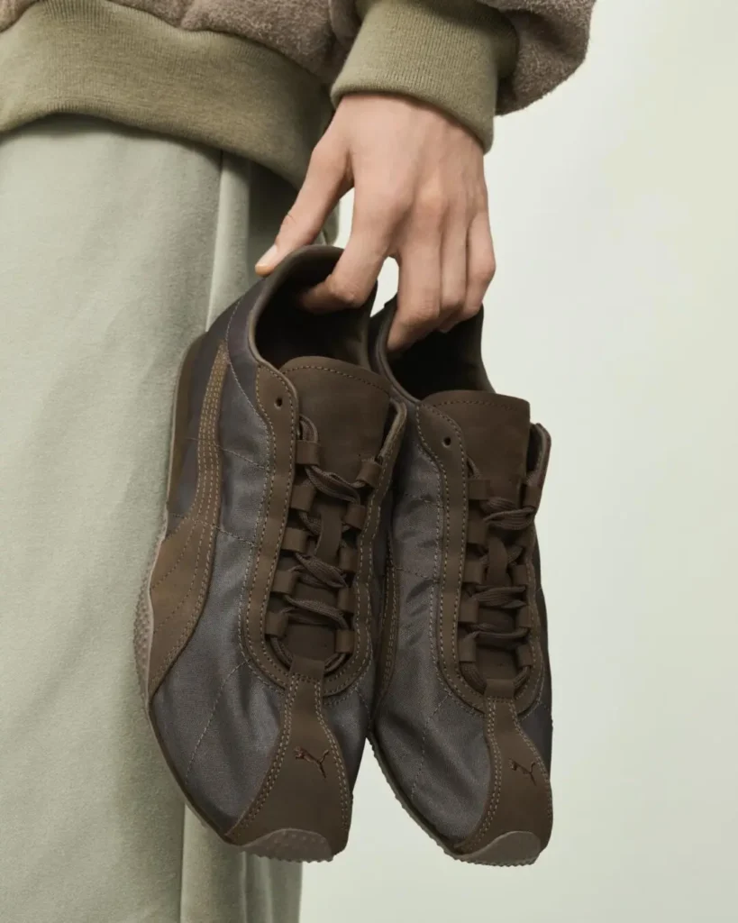 Hand holds a pair of brown and gray lace-up sneakers with visible Puma logos against a plain background
