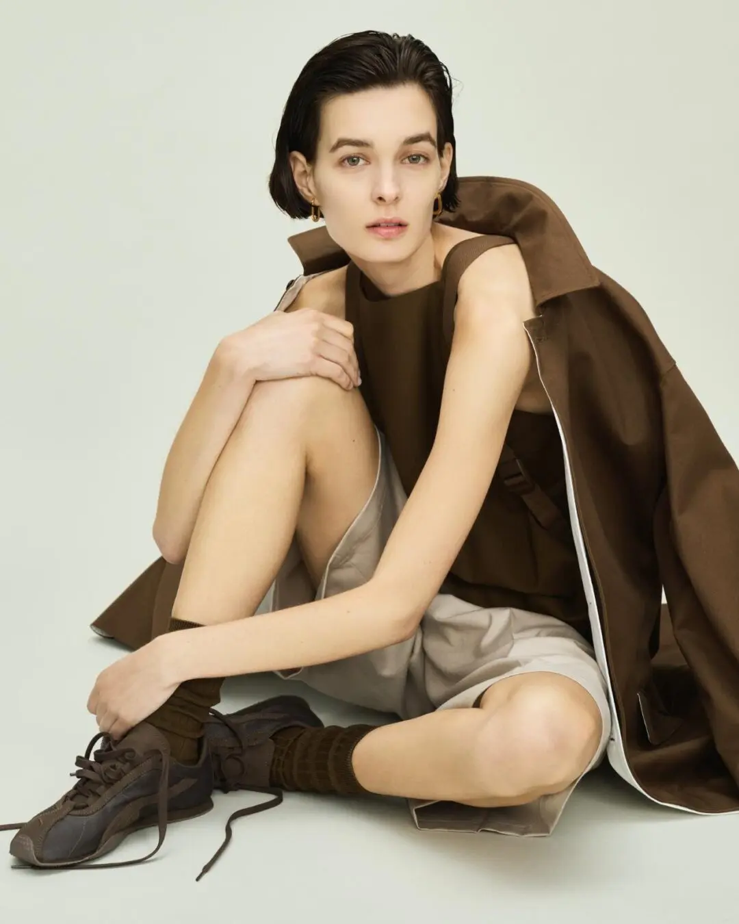Seated model in brown sleeveless top and jacket, gray shorts, socks, and brown lace-up sneakers against a pale backdrop