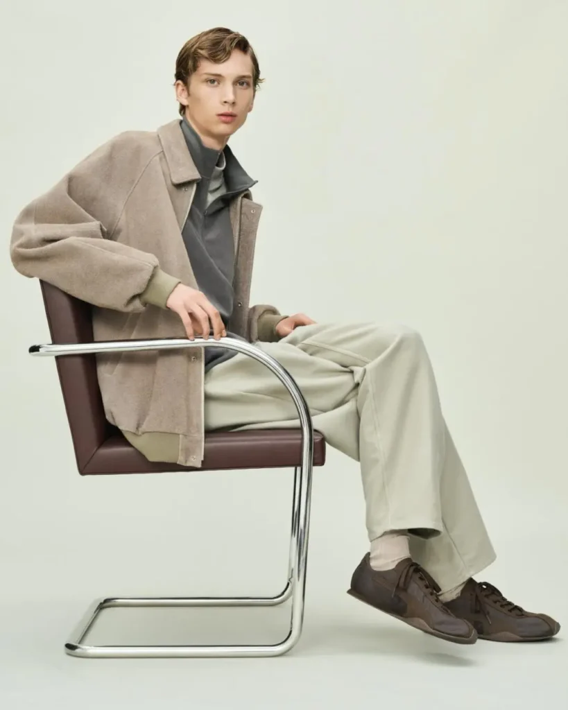 Young man seated in a brown leather-and-chrome chair, wearing a beige jacket, gray top, cream trousers, and brown shoes