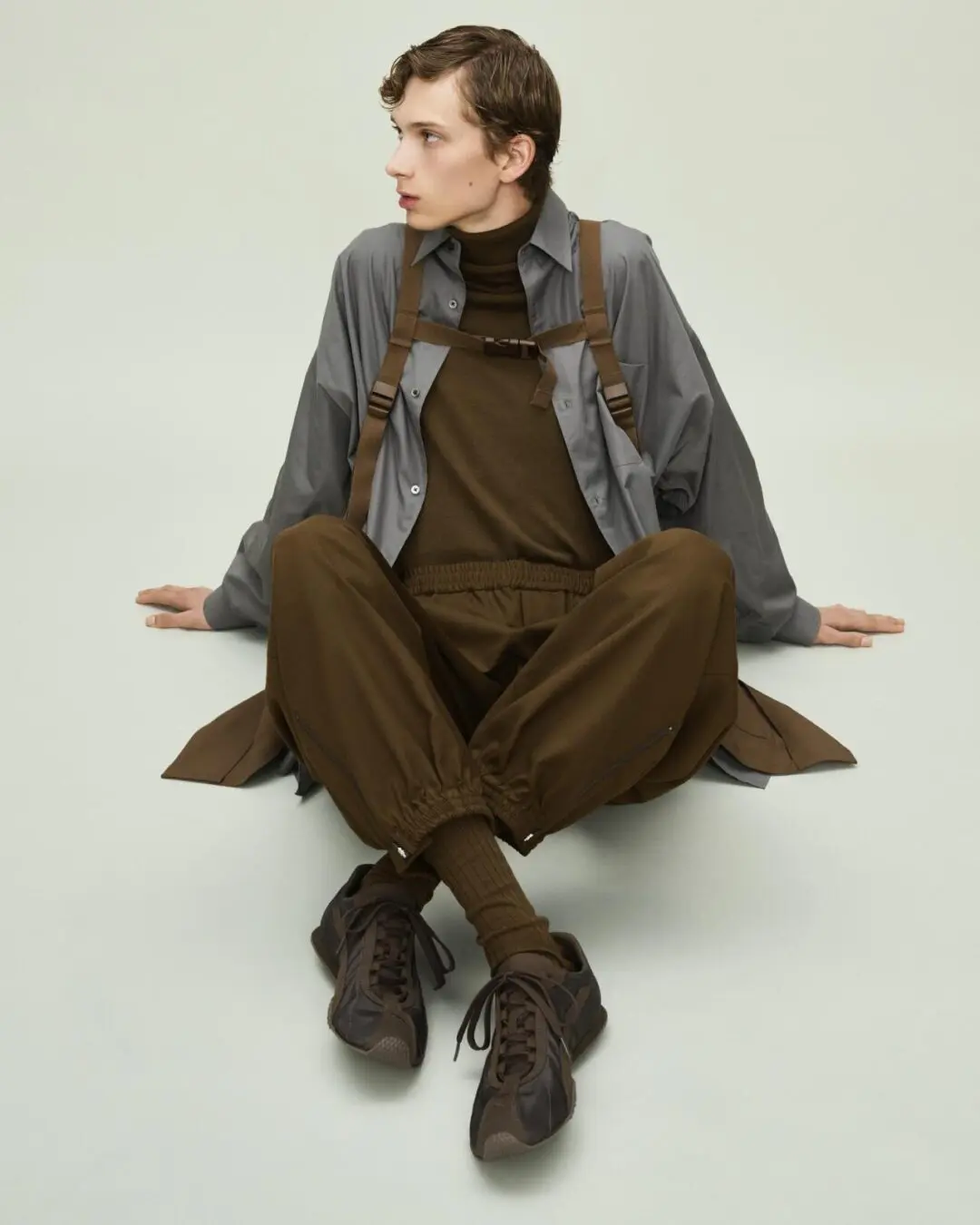 Seated model in brown layered clothing and harness, gray overshirt, and dark brown lace-up sneakers against a pale backdrop