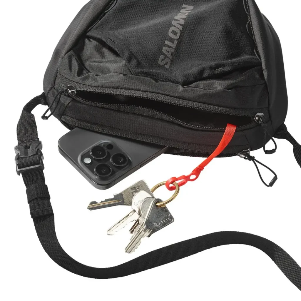 The Salomon shoulder bag shown carrying everyday essentials, including a smartphone and keys secured with the integrated red key clip inside the front zip pocket, demonstrating its practical organization and compact capacity