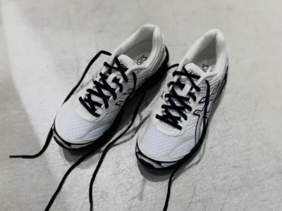 Pair of white and cream ASICS GEL-CUMULUS 16 running shoe with black laces resting on a polished concrete floor, shown with loosely untied lace