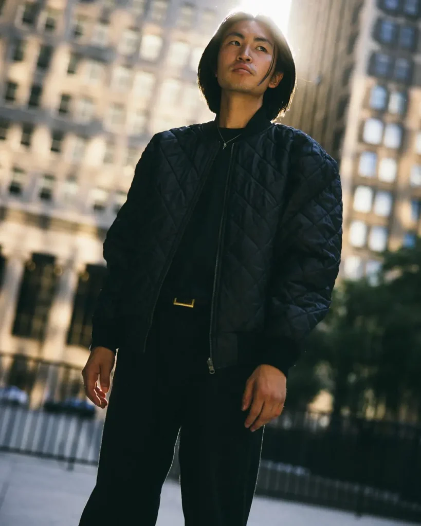 Person wear a black quilted bomber jacket stands in front of city buildings with warm afternoon light