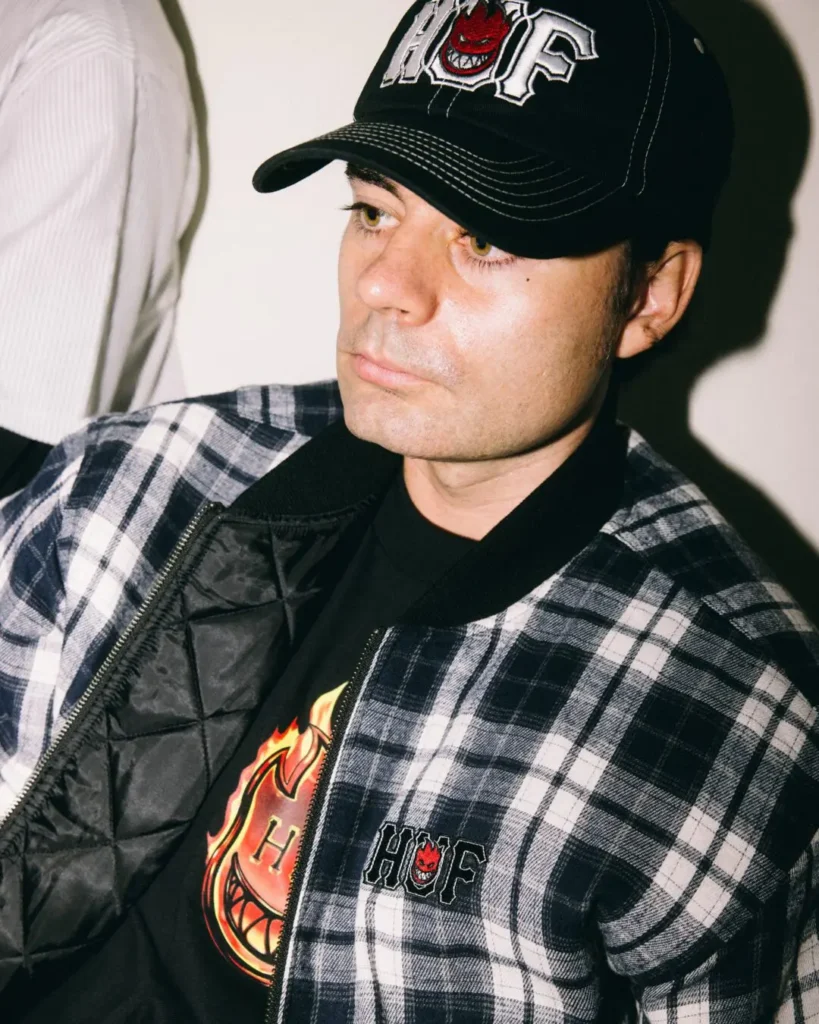 Close-up of a black plaid HUF jacket layered over a graphic Tee and paired with a HUF x Spitfire cap
