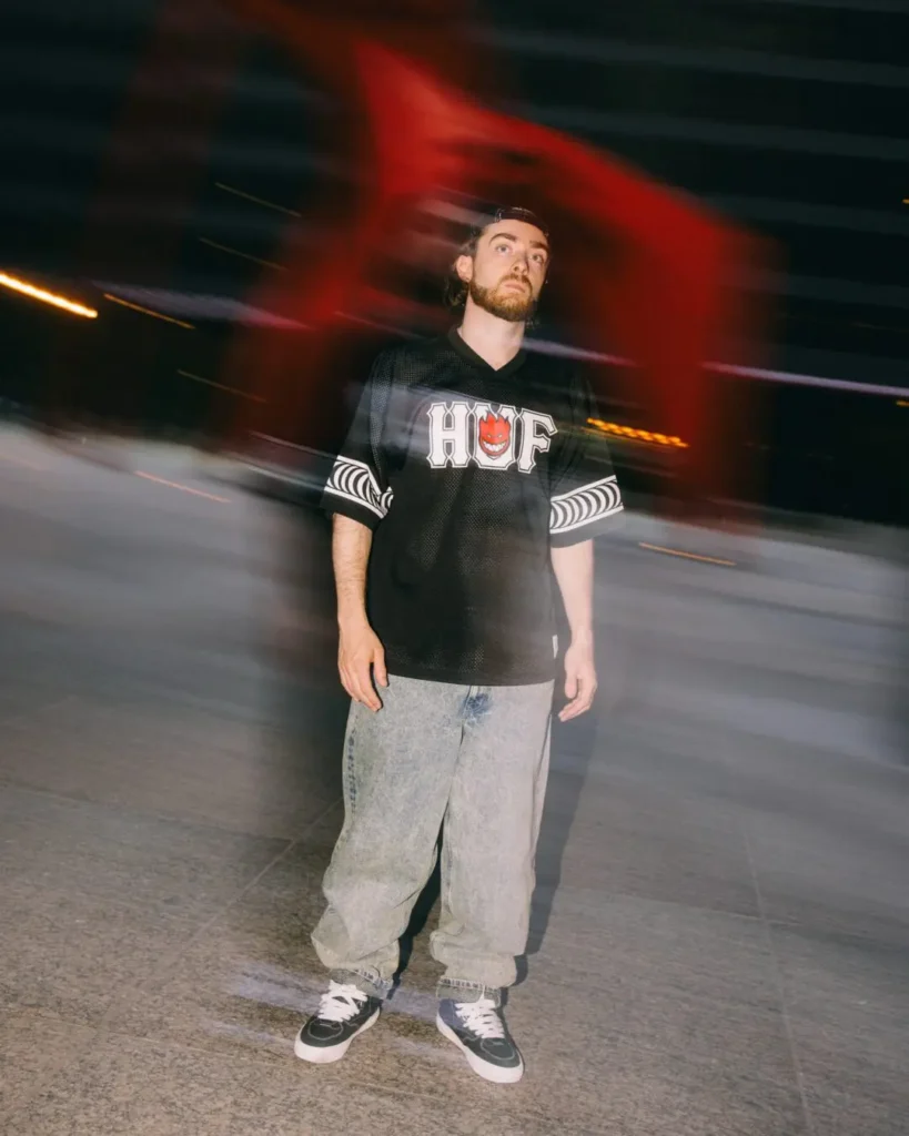 Person wearing a black HUF x Spitfire mesh jersey stands outdoors with motion-blurred city lights behind them