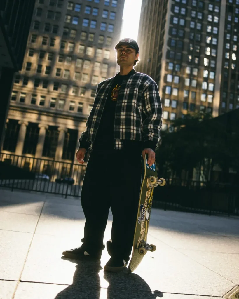Skateboarder holding a skateboard wears a plaid HUF jacket and graphic tee in a sunlit urban plaza