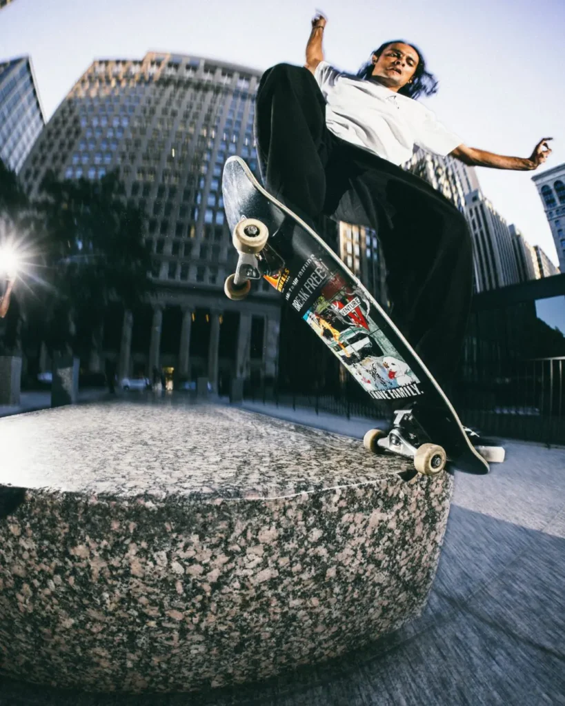 Skateboarder performs a trick over a granite ledge in a city plaza while wearing a white HUF Tee