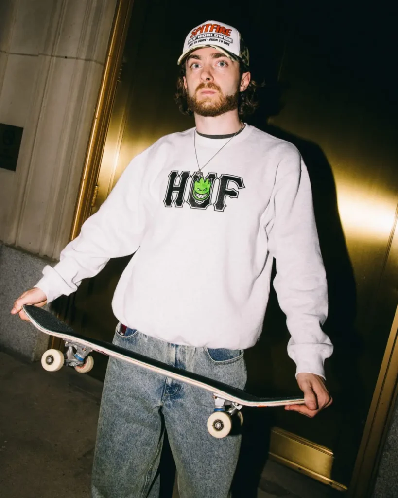Skateboarder holding a skateboard wears HUF and graphic tee crew neck sweatshirt in a sunlit urban plaza