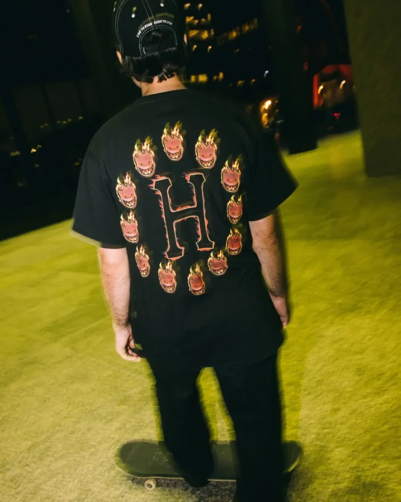 Rear view of a skateboarder wearing a black HUF x Spitfire graphic Tee featuring a large flaming "H" surrounded by Spitfire flame icons while riding through a city at night