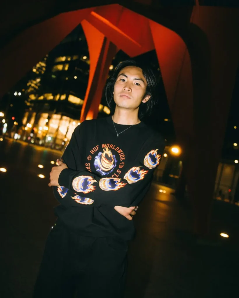 Person wearing a black HUF x Spitfire long-sleeve graphic shirt with flame graphics on the sleeves poses beneath a red architectural structure in a nighttime urban set
