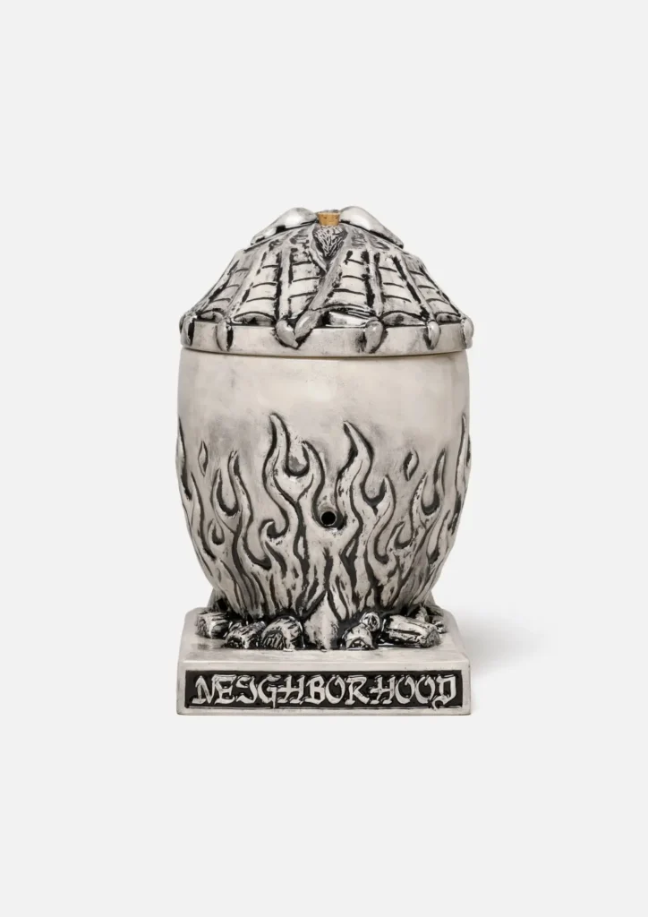 Rear view of the Neighborhood incense chamber. The rib textured back of the gargoyle lid is view. The body show the sculpt flame and a small circle smoke hole in the ceramic. The black plaque on the base is engrave with the word "NEIGHBORHOOD" in a white, gothic-style font