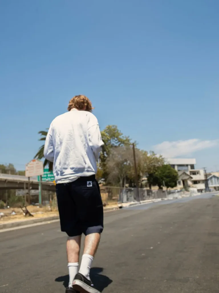Rear view of a model walking down a quiet street in an oversized white sweatshirt, dark knee-length shorts, crew socks, and black slip-on shoe