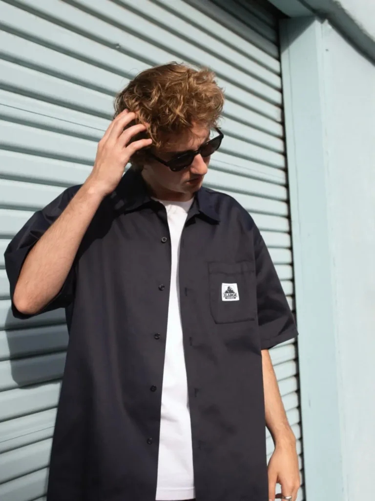 Model in a dark short-sleeve work shirt layered over a white Tee, paired with black sunglasses and standing against a light blue corrugated metal wall