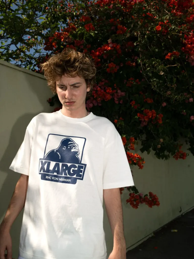 Model wearing a white graphic X-LARGE Tee featuring the signature gorilla logo, photographed outdoors in front of vibrant red flowering vine