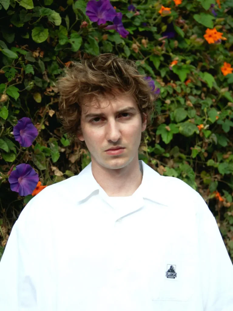 Portrait of a model in an oversized white short-sleeve shirt with a small X-LARGE chest logo, standing before lush green foliage and purple flower
