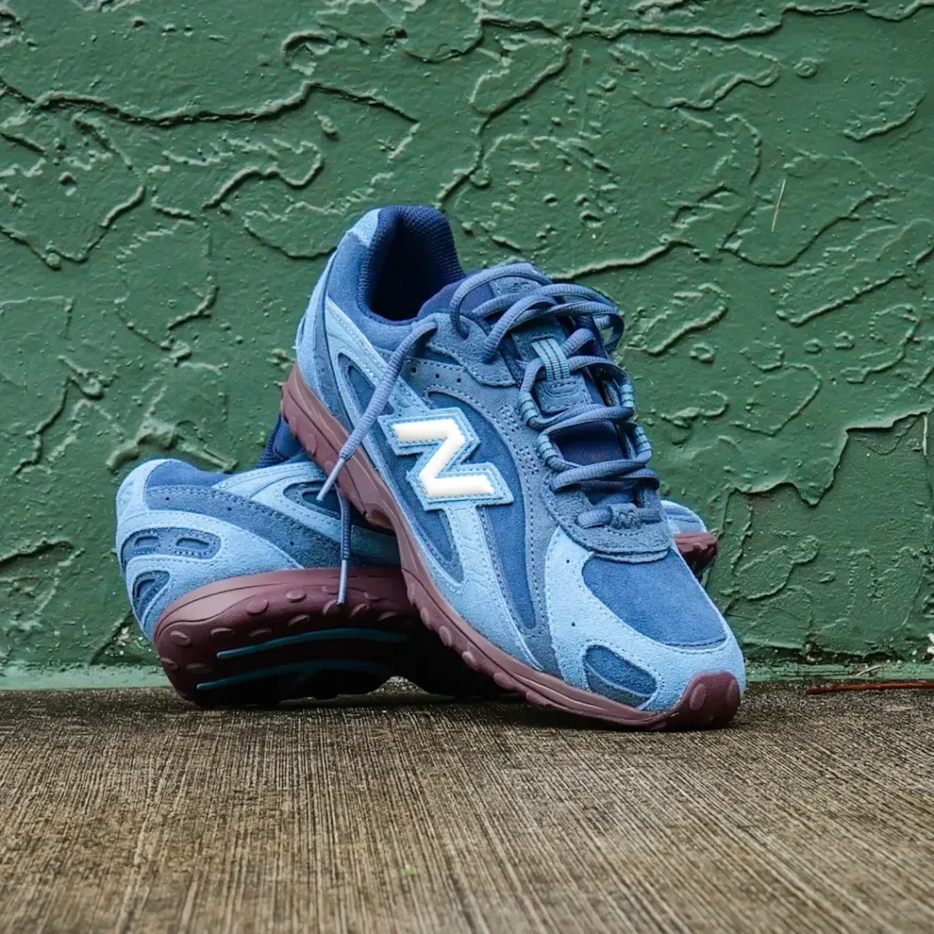 Blue New Balance shoes stacked against a textured green wall, featuring layered suede uppers, white “N” logos and burgundy sole