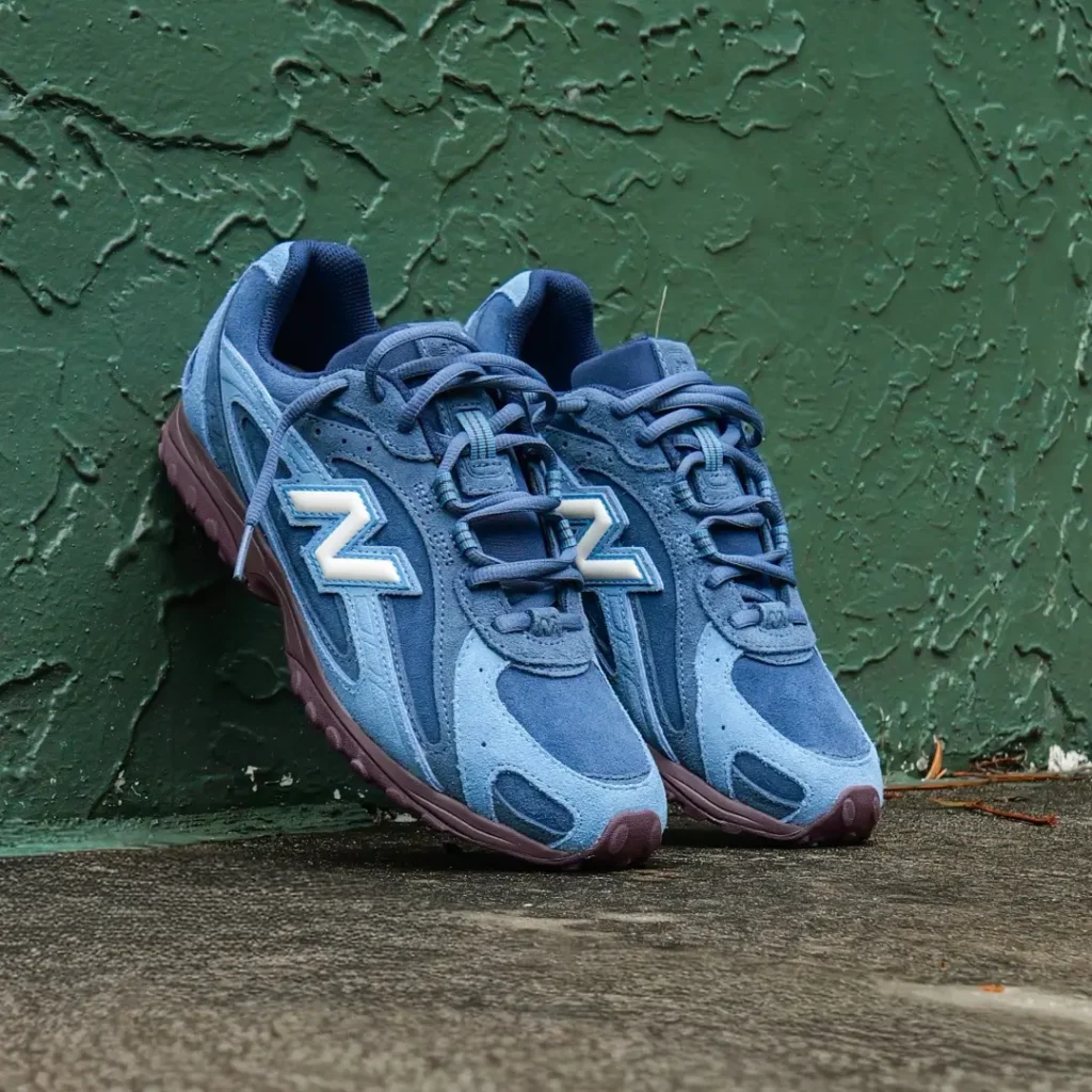 Pair of blue New Balance shoes leaning against a textured green wall, showing tonal suede panels, white “N” logos and burgundy sole