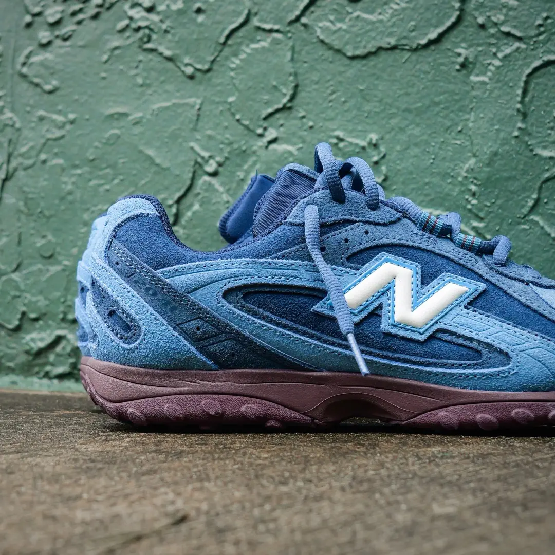 Close-up side view of a blue New Balance shoe featuring layered suede panels, a white N logo, tonal laces and a burgundy sole against a textured green wall.