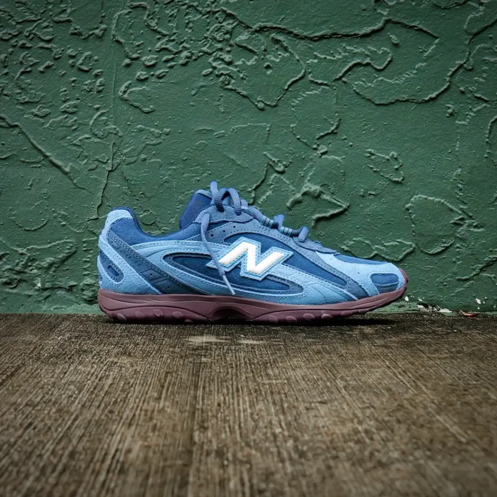 Side profile of a blue New Balance shoe position against a texture green wall with a burgundy sole and contrast white “N” logo
