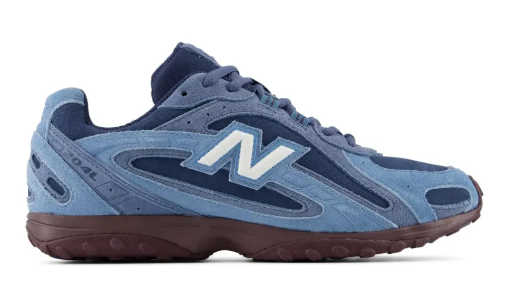 Product side view of a blue New Balance shoe on a white background, featuring tonal suede overlays, white “N” logo and burgundy sole