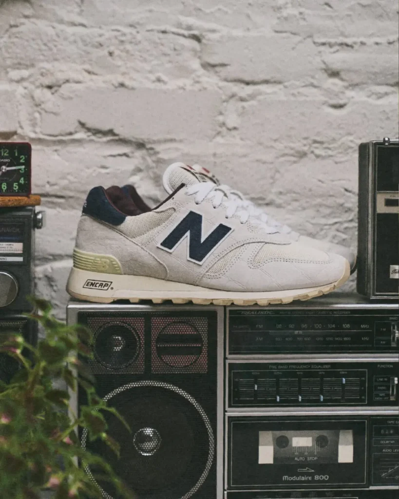 Side profile of the New Balance 1300 in off-white crocodile-textured suede with navy heel panel and N logo, rest atop a Real boombox stack beside a vintage alarm clock and houseplant