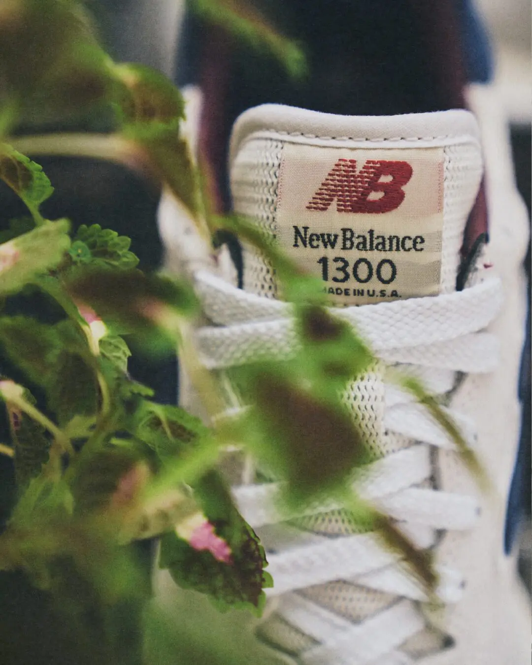 Close-up of the New Balance 1300 heel tab woven label reading New Balance 1300 Made in U.S.A., framed by white laces and green foliage in the foreground