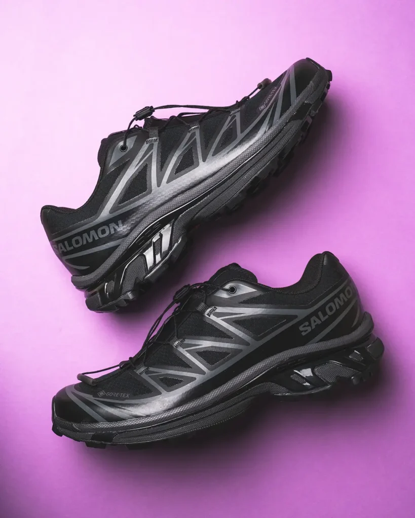 Two black and gray Salomon trail shoes with lugged soles and quick-lace toggles on a pink background