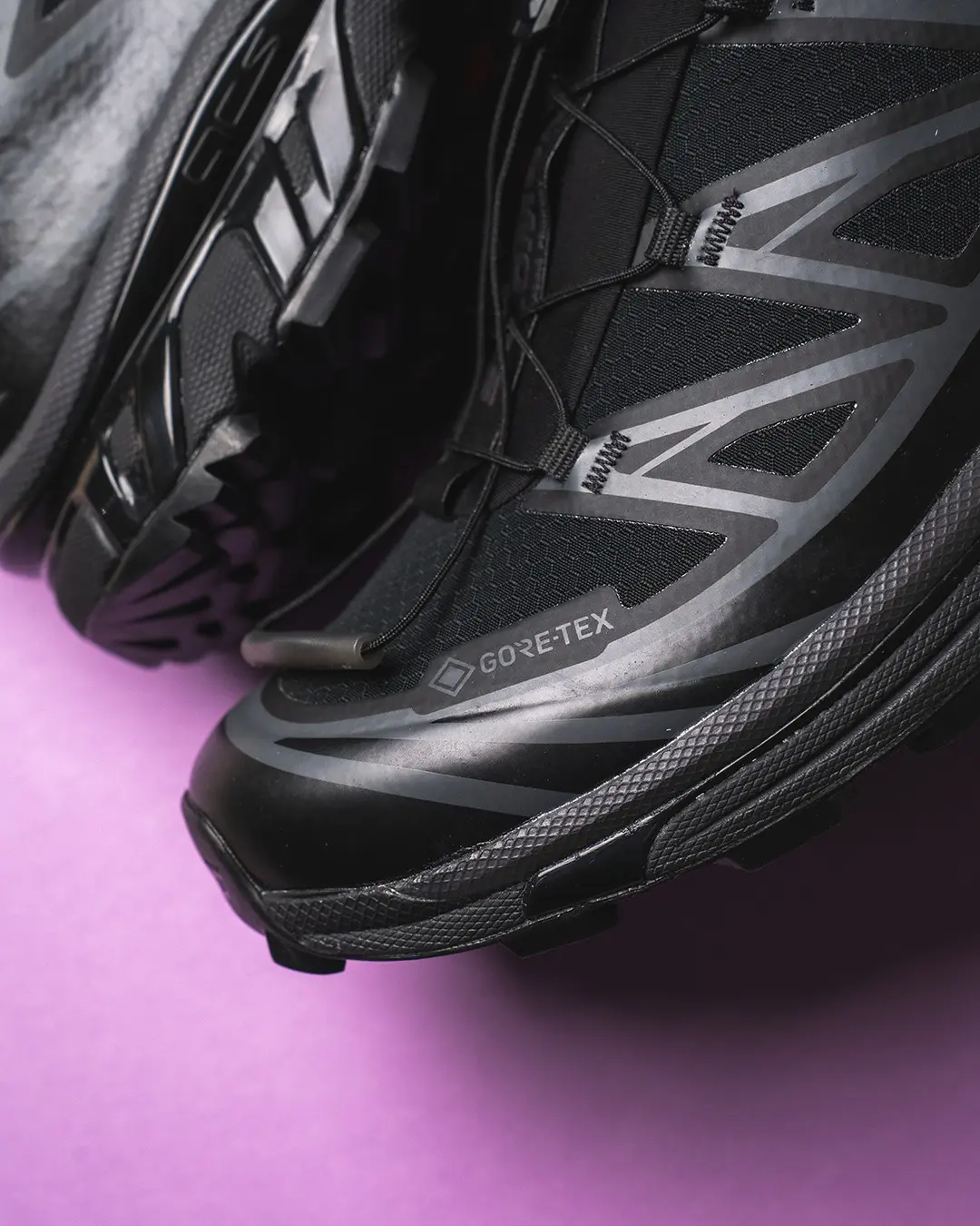Close-up of black Salomon trail shoes with gray overlays and visible GORE-TEX branding on a purple background